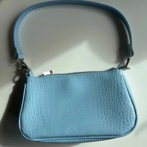 Ljusblå axelväska i krokopräglat skinn - Snygg ljusblå axelväska med krokodilpräglat mönster och silverfärgad dragkedja. Väskan har en stilren, avlång form och bred axelrem. Perfekt för att lyfta din outfit med en trendig touch.