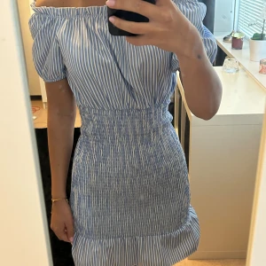 Blåvit offshoulder kortklänning - Säljer en söt blåvit randig kortklänning med offshoulder och puffärmar. Klänningen har smockad midja och volang nertill, vilket ger en feminin och somrig känsla. Perfekt för dig som gillar en fräsch och trendig stil.