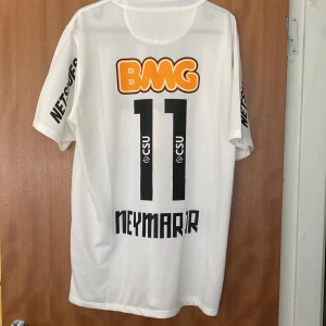 Vit Santos Neymar 11 - Helt oanvänd Santos hemmatröja från Nike med Neymar 11-tryck på ryggen. Vit med svarta och orangea detaljer, klubbmärke på bröstet och flera sponsorloggor. Tillverkad i Dri-Fit-material som andas, perfekt för fotbollsträning eller match.