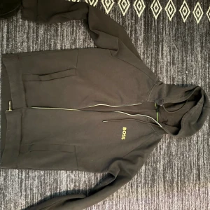 Svart hoodie från BOSS med dragkedja - Svart hoodie från BOSS med gul logga på bröstet, hel dragkedja och huva med snörning. Snygga gröna detaljer på insidan av kragen. Perfekt för dig som gillar stilrena och bekväma plagg.