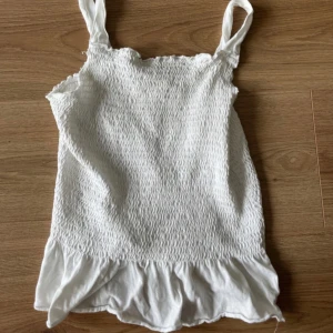 Vit smockad linnetopp från Zara Kids - Supersöt vit linnetopp från Zara Kids med smockad överdel och volangkant nertill. Tunt och mjukt bomullstyg, perfekta breda axelband och en luftig känsla. Enkel att matcha till jeans eller kjol för en fräsch sommarlook. Passar någon som har strl. XS