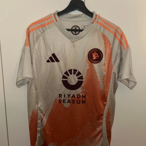 Adidas Riyadh Season fotbollströja M - Snygg fotbollströja från Adidas med Riyadh Season-tryck. Tröjan är ljusgrå med orange detaljer och har tre klassiska ränder på axlarna. Unik design med orange mönster framtill och rund hals. Perfekt för dig som gillar sportig och modern stil.