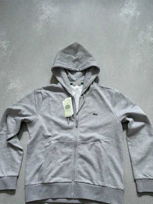 Lacoste zip up - Classic Lacoste zip-up jacket in a versatile grey colorway, featuring the iconic embroidered crocodile logo on the chest. A timeless piece that blends sporty and casual style, perfect for everyday wear or layering.