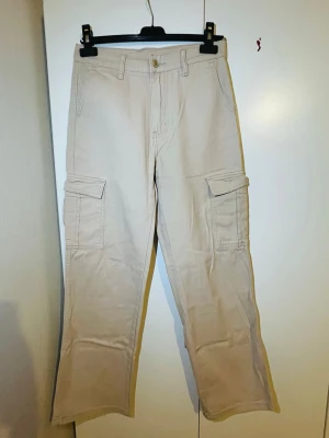 Beige cargopants med raka ben - Säljer ett par beige cargopants med raka ben och stora fickor på sidorna. Byxorna har normal midja, bälteshällor och stängs med knapp och dragkedja. Tillverkade i bomull för en skön och avslappnad känsla.