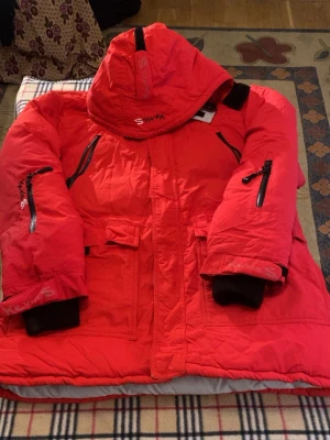 Röd parkas från Kevin S. XL - Säljer en röd parkas från Kevin S. i storlek XL. Jackan har huva, flera fickor med dragkedjor och muddar vid ärmsluten. Perfekt för kalla dagar och har coola detaljer med loggan på både ärm och rygg. Materialet är tåligt och känns riktigt robust.