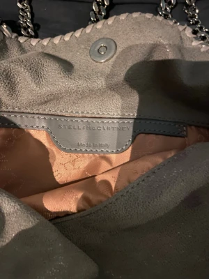 Grå axelväska Stella McCartney - Säljer en snygg grå axelväska från Stella McCartney med kedjedetaljer runt kanterna och lång axelrem i metall. Väskan är tillverkad i syntetmaterial och har en mjuk, mockaliknande yta. Insidan är fodrad i beige tyg och väskan stängs med magnetknapp.