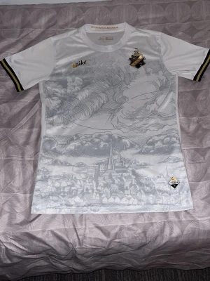 Vit AIK x Nike fotbollströja - Säljer en vit AIK x Nike fotbollströja med snyggt stadsmotiv i grått på framsidan. Tröjan har svarta och gula detaljer på ärmsluten och AIK-emblem på bröstet. Perfekt för dig som vill sticka ut på läktaren eller i stan.