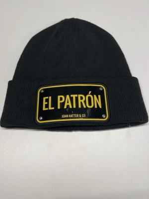 Svart mössa El Patrón John Hatter & Co - Säljer en svart ribbstickad mössa från John Hatter & Co med en stor metallbricka framtill där det står 'EL PATRÓN' i gult. Mössan har uppvikt kant och är riktigt clean och stilren, perfekt för dig som vill sticka ut. Den är helt oanvänd.
