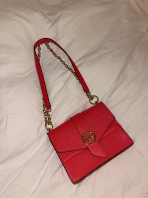 Axelremsväska från Michael Kors  - Säljer en snygg korallfärgad axelväska från Michael Kors med guldfärgade kedjedetaljer och logga framtill. Väskan är aldrig använd och har inget slitage någonstans - inte ens på kedjan/loggan!!!