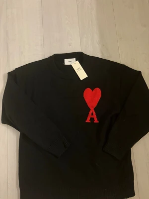Svart stickad tröja från Ami Paris  - Svart stickad tröja från Ami med rött hjärta och bokstaven A broderat på bröstet. Klassisk rund halsringning och långa ärmar. Perfekt statement-plagg för dig som gillar stilrena detaljer och vill ha något som sticker ut.