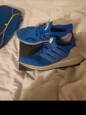 Blå Adidas Ultraboost sneakers - Säljer ett par blå Adidas Ultraboost sneakers med vit och svart sula. Ovandelen är i ventilerande syntetmaterial och har snörning. Skorna har en klassisk rund tå och platt sula, perfekt för dig som gillar sportig stil och komfort.Kom gärna med prisförslag också 