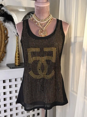 Svart glittrig linnetopp med nitar - Säljer en svart linnetopp med glittriga guldfärgade och kopparfärgade nitar i ett rutmönster och stor logga framtill. Toppen är ärmlös och har en loose passform med rund halsringning. Materialet är tunt och lätt, perfekt för att sticka ut.