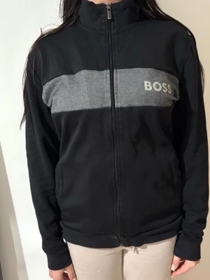 Svart ziptröja från BOSS - Säljer en svart ziptröja från BOSS med grå panel och logga över bröstet. Tröjan har hög krage, hel dragkedja och långa ärmar. Perfekt för en chill och stilren look.