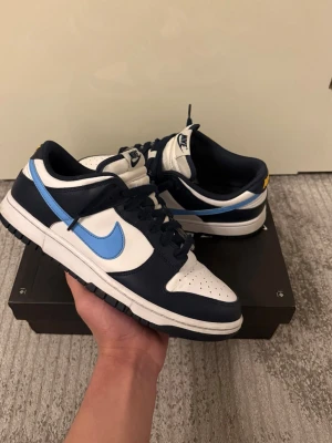 Nike Dunk Low blå och vita sneakers - Säljer ett par Nike Dunk Low sneakers i marinblått och vitt med ljusblå swoosh och gula detaljer på hälen. Skorna har klassisk låg siluett, perforerad tåbox och snörning. Ovandelen är i skinn och de har en vit mellansula med svart yttersula. Perfekta för dig som gillar streetwear.