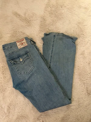 True Religion Joey ljusblå flare jeans - Säljer ett par True Religion Joey jeans i ljusblå tvätt med klassiska kontrastsömmar och ikonisk fickdetalj bak. Modellen är low rise med flare-ben och tillverkad i mjukt jeansmaterial. Perfekta för dig som gillar retrovibbar och vill sticka ut lite extra. Säljer pga att de är förstora. Liten fläck längst ner på benet, går säkert bort i tvätt, kan absolut tvätta om så önskas.