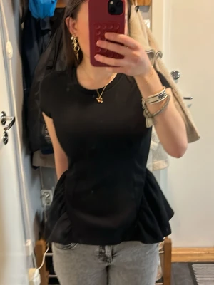 Svart peplum topp  - Säljer en svart peplum topp med rund halsringning och korta ärmar. Toppen har en figurnära passform upptill och volangliknande utställd nederdel som ger en snygg siluett. 