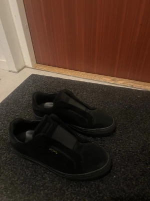 Svarta sneakers från Axel Arigato - Säljer ett par svarta sneakers från Axel Arigato i mocka med minimalistisk design och diskret guldfärgad logga på sidan. Skorna har platt sula, rund tå och elastisk detalj över vristen för enkel påtagning. Perfekta för en clean och stilren look. Helt nya, använts en enda gång och de kommer med box 