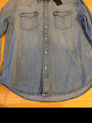 Levi's blå jeansskjorta - Klassisk blå jeansskjorta från Levi's med tryckknappar och två bröstfickor med lock. Skjortan har lång ärm och western-inspirerade sömmar. Perfekt till ett avslappnat streetwear-utseende.