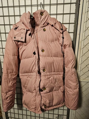 Rosa dunjacka från Boomerang XXS - Säljer en fluffig rosa dunjacka från Boomerang i storlek XXS. Jackan har huva, stora knappar, dragkedja och snörning vid huvan. Yttermaterial i polyester och fyllning av 90% dun och 10% fjäder. Perfekt för kalla dagar och riktigt mysig att bära.