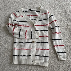 Vit randig kofta från JC - Säljer en vit kofta från JC i storlek XS med tunna svarta och röda ränder. Koftan har v-ringning, ribbade muddar och knäppning med två knappar framtill. Perfekt för lager på lager och enkel att styla till olika outfits. Obs: en liten fläck längst nere på koftan, se bild 