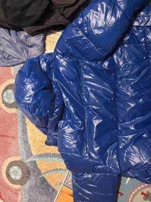 Blå pufferjacka från Pull&Bear - Säljer en blå pufferjacka från Pull&Bear i storlek L. Jackan har huva, dragkedja och en glansig finish. Perfekt för kalla dagar och riktigt skön att bära. Enkel och stilren design som funkar till det mesta.