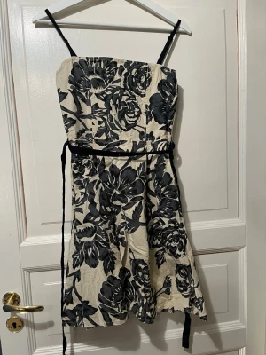 Blommig kortklänning med smala band - Snygg kort klänning med svart-vitt blommönster och smala axelband. Klänningen har ett band i midjan som kan knytas för extra form. Perfekt för dig som gillar en feminin och trendig stil.
