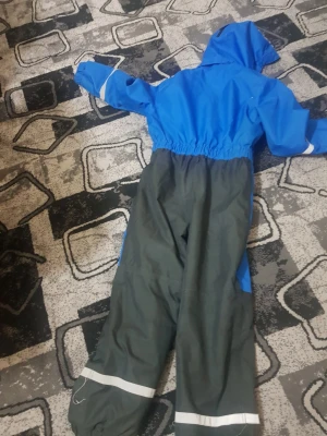 Blå täckbyxa overall Graffiti - Blå och mörkgrå täckbyxa overall från Graffiti med huva och resår i midjan. Overallen har reflexdetaljer på ärmar och ben, samt dragkedja framtill. Materialet är tåligt och passar perfekt för kalla dagar utomhus.