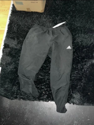 Svarta träningsbyxor från Adidas - Svarta träningsbyxor från Adidas med klassisk logga på låret. Byxorna har resår i midjan och är gjorda i ett lätt och syntetiskt material som passar perfekt för sport och träning. Enkel och stilren design som är lätt att matcha.