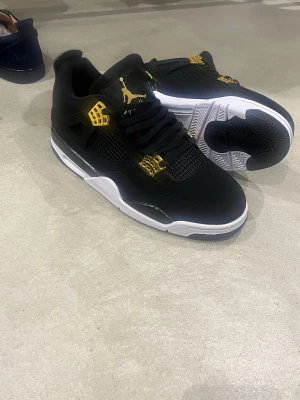 Nike Air Jordan 4 svart/guld - Säljer ett par Nike Air Jordan 4 sneakers i svart med snygga gulddetaljer och vit sula. Skorna har klassisk snörning, meshpaneler och Jumpman-logga på plös och häl. Perfekta för dig som vill sticka ut med stilrena och ikoniska sneakers.