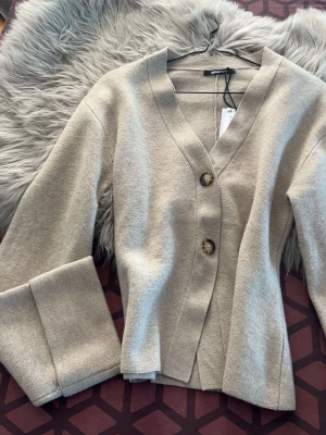 Beige kofta från Gina Tricot - Stilren beige kofta från Gina Tricot med två stora knappar framtill. Koftan har v-ringad hals, långa ärmar och är gjord i ett mjukt stickat material. Perfekt för lager på lager och enkel att matcha med olika outfits. Har aldrig tagit på mig säljer pga fel storlek köptes för 460