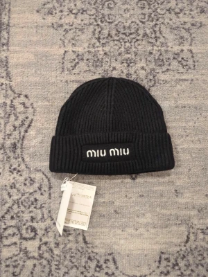 Svart ribbstickad mössa från Miu Miu - Säljer en svart ribbstickad mössa från Miu Miu med uppvikt kant och broderad logga framtill. Mössan är tillverkad i 100% ull och har en klassisk, stilren look som passar perfekt till kyliga dagar.