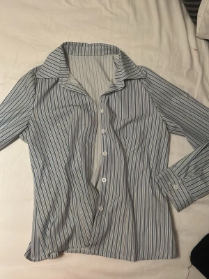 Randig blåvit skjorta med knappar - Snygg skjorta med blå och vita ränder, klassisk krage och knäppning framtill. Långärmad modell som passar perfekt till jeans eller kjol. Skjortan har en avslappnad passform och är tillverkad i ett mjukt bomullsliknande material.