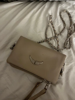 Beige axelväska Zadig & Voltaire - Snygg beige axelväska från Zadig & Voltaire i slätt skinn. Väskan har silverfärgad kedjerem och ett emblem med vingar framtill. Insidan är fodrad med svart tyg med logga. Perfekt storlek för det viktigaste och har dragkedja samt tofsdetaljer. Liten defekt kolla bild två 