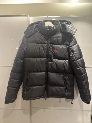 Svart pufferjacka från Polo Ralph Lauren - Säljer en svart pufferjacka från Polo Ralph Lauren med klassisk röd logga på bröstet. Jackan har hög krage, avtagbar huva och flera dragkedjeförsedda fickor. Perfekt för kalla dagar och riktigt snygg till alla outfits.
