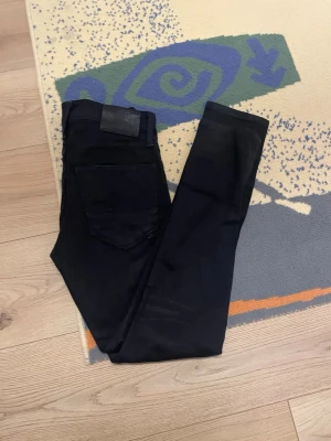 Svarta jeans från Jack & Jones - Säljer ett par svarta jeans från Jack & Jones med klassisk femficksdesign och svart läderpatch bak i midjan. Jeansen har rak passform och är tillverkade i ett mjukt bomullsmaterial med lite stretch för extra komfort.