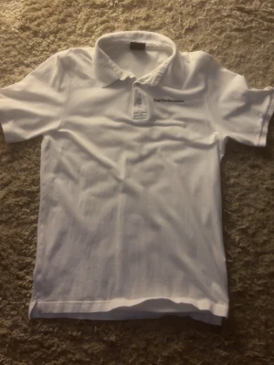 Vit Peak Performance pikétröja - Klassisk vit pikétröja från Peak Performance i bomull. Tröjan har korta ärmar, krage och knappar framtill samt diskret logga broderad på bröstet. Perfekt till en clean och sportig look.