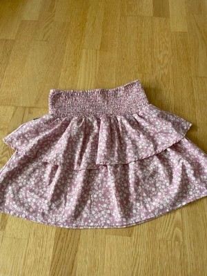 Rosa blommig volangkjol - Supersöt somrig rosa kortkjol med vita små blommor och bred smock i midjan. Storlek xs eller s men passar många storlekar då den är väldigt stretchig. 