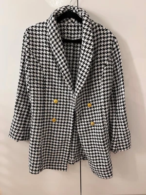 Svartvit rutig kavaj med guldknappar - Säljer en snygg svartvit kavaj med klassiskt hundtandsmönster och fyra guldiga knappar framtill. Kavajen har bred krage och är i ett mjukt, vävt material som känns lyxigt. 