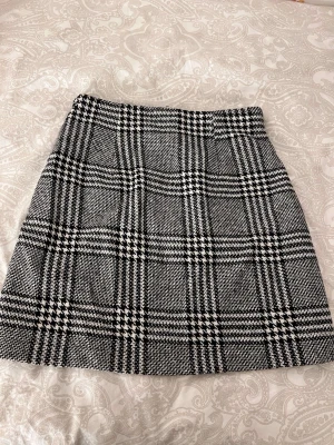 Rutig svartvit kortkjol med dragkedjor - Snygg kortkjol i svartvitt rutigt mönster med två dragkedjefickor framtill och en dragkedja mitt fram. Kjolen har en klassisk A-linjeform och känns både trendig och tidlös. 