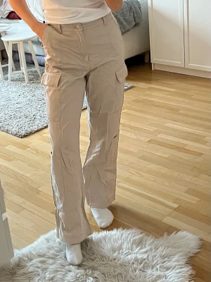 Beige cargopants med raka ben - Säljer ett par beige cargopants med raka ben och stora fickor på sidorna. Byxorna har hög midja och är tillverkade i ett mjukt bomullsmaterial. Perfekta för en chill och avslappnad stil. Passformen är loose och de har klassiska cargodetaljer.