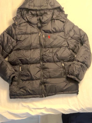 Svart pufferjacka från Polo Ralph Lauren - Säljer en svart pufferjacka från Polo Ralph Lauren i storlek L. Jackan har huva, flera dragkedjefickor och klassisk röd logga på bröstet. Den är quiltad och har en riktigt skön och varm känsla, perfekt för kyliga dagar. Perfekt om du söker en mer Exklsuiv stil, passar perfekt till skolan eller finare tillfällen 