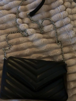 Svart quiltad axelväska med kedja - Säljer en svart axelväska i quiltad design med snyggt V-mönster. Väskan har en lång silverfärgad kedjerem med en liten skinnbit upptill för extra komfort. Perfekt storlek för det viktigaste och ger en cool vibe till din outfit. 13cm x 23cm ish. Ganska lång kedja