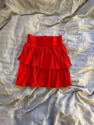 Röd volangkjol med resårmidja - Supersöt röd kortkjol med bred resår i midjan och två lager volanger. Kjolen har en plisserad struktur som ger extra rörelse och volym. Perfekt för dig som vill sticka ut med en färgstark och lekfull stil.