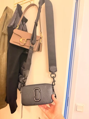 Svart axelväska från Marc Jacobs - Säljer en svart axelväska från Marc Jacobs med bred justerbar axelrem och dubbla dragkedjor. Väskan är i skinn med präglad logga framtill och har en stilren, boxig form. Superfint skick, pris kan diskuteras 😊