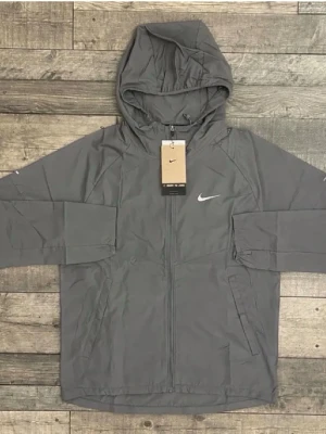 Grå vindjacka från Nike med huva - Snygg grå vindjacka från Nike med huva och dragkedja framtill. Jackan har en minimalistisk look med diskret Nike-logga på bröstet och praktiska sidofickor. Tillverkad i lätt polyester som passar perfekt för blåsiga dagar.