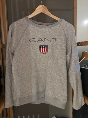 Grå sweatshirt från GANT - Säljer en grå sweatshirt från GANT med broderad logga och sköld på bröstet. Tröjan har rund halsringning och långa ärmar. Perfekt för dig som gillar klassisk och sportig stil.