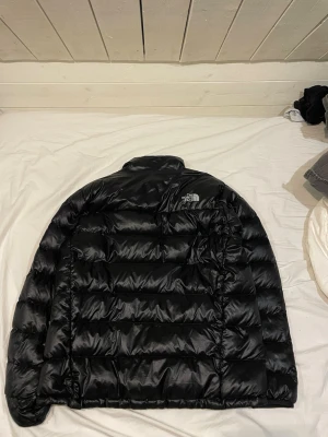 Svart pufferjacka från The North Face - Säljer en svart pufferjacka från The North Face med hög krage och diskret logga på bröstet. Jackan har en quiltad design och dragkedja framtill. Perfekt för kalla dagar och riktigt snygg till streetwear-looken.