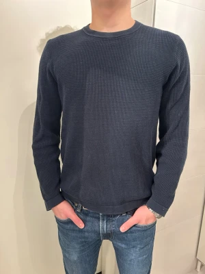 Mörkblå stickad tröja  - Mörkblå långärmad tröja från Selected Homme i ett strukturerat mönster. Tröjan har rund halsringning och en normal passform. Perfekt för lager-på-lager under vinter och vår. Snygg och enkel att matcha med jeans! Storlek är S, och modellen är 176cm. 