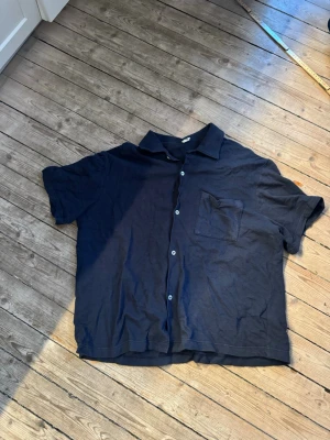 Mörkblå kortärmad skjorta H&M - Snygg mörkblå skjorta från H&M med korta ärmar och klassisk krage. Skjortan har knappar framtill och en bröstficka på vänster sida. Perfekt till sommaren och enkel att matcha med olika outfits.