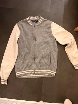 Gråvit bomberjacka från Primark - Säljer en stilren bomberjacka från Primark i grått med vita ärmar. Jackan har dragkedja framtill, ribbade muddar och krage med vita och grå ränder. Perfekt för dig som gillar en sportig look.